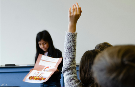 Ein Kind steht vor einer Gruppe anderer Kinder im klassenzimmer und hält ein Poster in der Hand. Im Vordergrund meldet sich ein Kind.