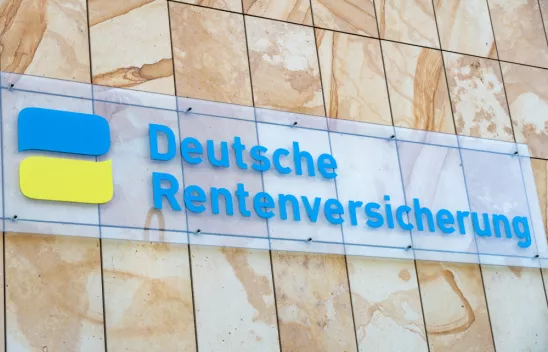 An einer hellen Steinwand hängt ein Glasschild mit der blauen Aufschrift "Deutsche Rentenversicherung". Links neben der Schrift ist ein Zeichen in Form von zwei Sprechblasen in gelb und blau. An einer hellen Steinwand hängt ein Glasschild mit der blauen Aufschrift "Deutsche Rentenversicherung". Links neben der Schrift ist ein Zeichen in Form von zwei Sprechblasen in gelb und blau.