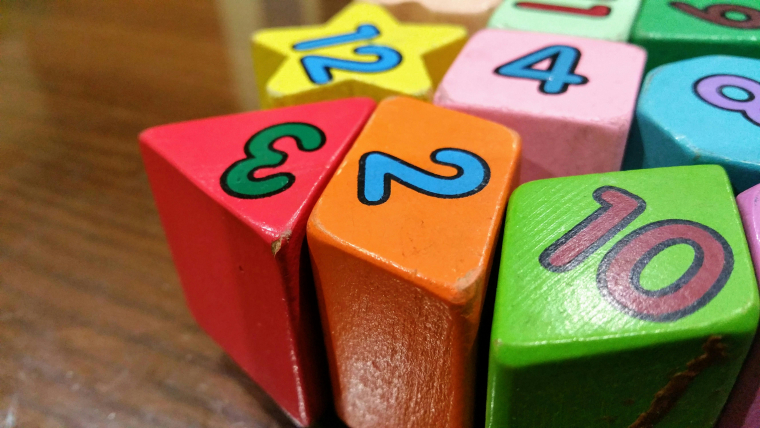 Bunte Spiel-Bauklötze in den Farben rot, orange, grün, rosa, hellblau und gelb. Darauf stehen jeweils Zahlen. 