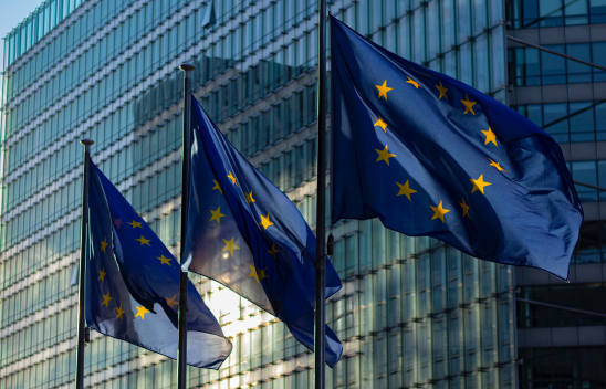 Drei europäische Flaggen (blauer Hintergrund mit einem gelben Sternenkreis) wehen vor einem hohen Glasgebäude.