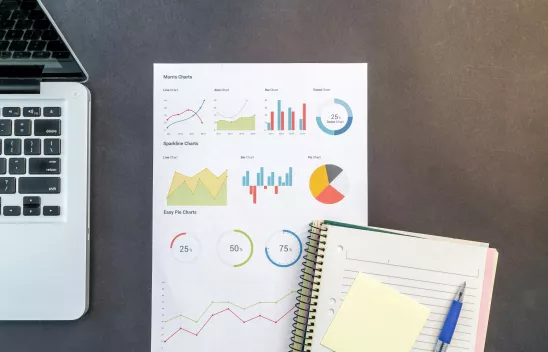 Arbeitsfläche mit einem geöffneten Laptop, einem Blatt mit verschiedenen bunten Diagrammen und einem Notizbuch