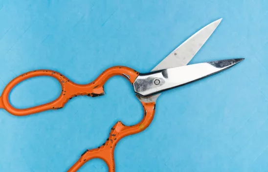 Eine geöffnete Schere mit orangefarbenen Griff liegt auf einer blauen Fläche. Eine geöffnete Schere mit orangefarbenen Griff liegt auf einer blauen Fläche.