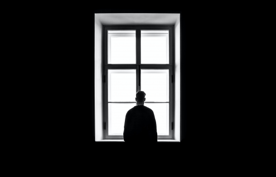 Eine Schwarz-Weiß-Aufnahme einer Person, die von hinten fotografiert wurde und in einem dunklen Raum alleine vor einem Fenster steht und hinaufblickt. 