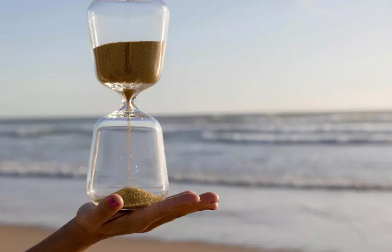 Eine Hand hält eine Sanduhr. Im Hintergrund sieht man Strand und Meer.