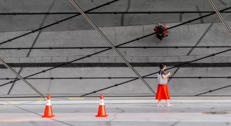 Eine Person mit rotem Rock läuft vor einer Glasfassade, auf der sich die Struktur des Straßenbelags und rote Baustellenkegel spiegeln.