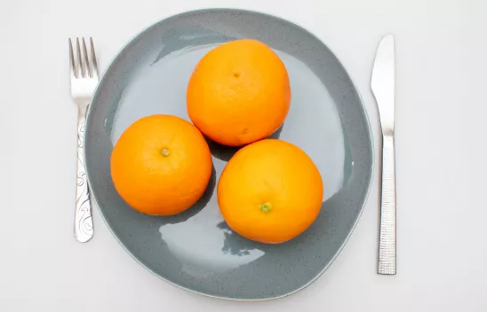 Drei Orangen auf einem Teller, rechts und links davon liegen Messer und Gabel.
