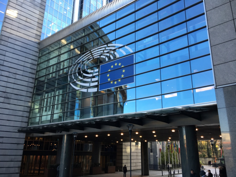 Ein Gebäude mit einer Glasfront, auf der mittig ein Konferenzsaal in weiß und die europäische Flagge (blauer Hintergrund mit einem gelben Sternenkreis) aufgeklebt sind.