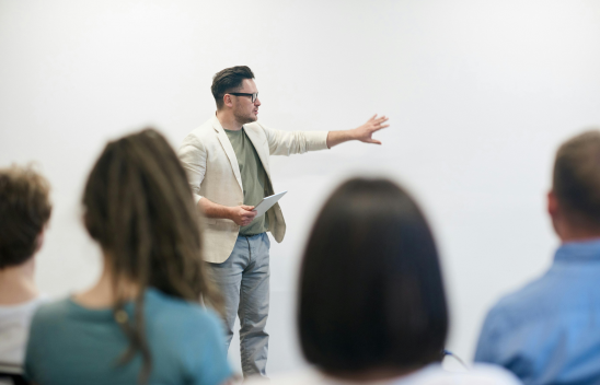 Person hält einen Vortrag vor einer Gruppe und gestikuliert in Richtung einer weißen Wand