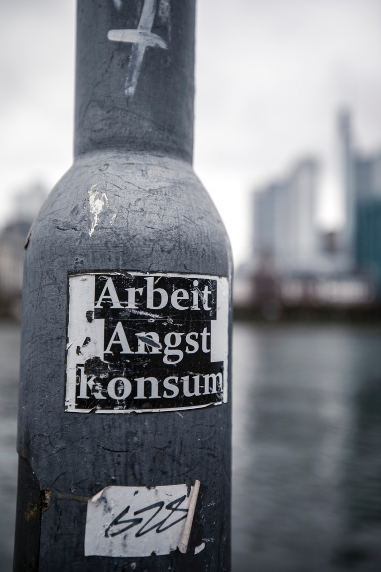 Ein zerkratzter Sticker mit der Aufschrift "Arbeit, Angst, Konsum" klebt an einer Laterne. Das Foto ist Schwarzweiß. 