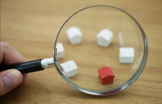 Eine Lupe wird von einer Hand gehalten und zu sehen sind fünf kleine weiße Holzhäuser sowie ein rotes kleines Holzhaus