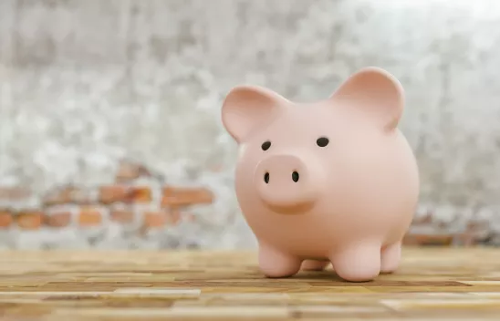 Ein rosafarbenes Sparschwein steht auf einem Holzboden vor einer Steinwand. Ein rosafarbenes Sparschwein steht auf einem Holzboden vor einer Steinwand.