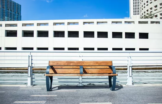 Foto einer Holzbank am Straßenrand. In der Mitte der Bank ist eine dritte Lehne, die das Liegen auf der Bank unmöglich macht und ein Beispiel für wohnungslosenfeindliche Architektur ist. Foto einer Holzbank am Straßenrand. In der Mitte der Bank ist eine dritte Lehne, die das Liegen auf der Bank unmöglich macht und ein Beispiel für wohnungslosenfeindliche Architektur ist.