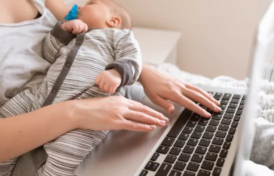 Eine vielbeschäftigte junge Frau arbeitet oder lernt zuhause an ihrem Notebook, während sie ihr Baby auf dem Arm hält Eine vielbeschäftigte junge Frau arbeitet oder lernt zuhause an ihrem Notebook, während sie ihr Baby auf dem Arm hält
