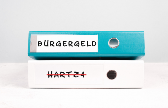 Ein blauer Ringordner mit der seitlichen Aufschrift "Bürgergeld" liegt auf einem weißen Ringordner mit der in rot durchgestrichenen Aufschrift "Hartz 4".