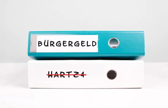 Ein blauer Ringordner mit der seitlichen Aufschrift "Bürgergeld" liegt auf einem weißen Ringordner mit der in rot durchgestrichenen Aufschrift "Hartz 4". Ein blauer Ringordner mit der seitlichen Aufschrift "Bürgergeld" liegt auf einem weißen Ringordner mit der in rot durchgestrichenen Aufschrift "Hartz 4".