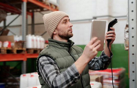 Person mit Mütze und Weste scannt Ware in einem Lager mit Tablet und Handscanner