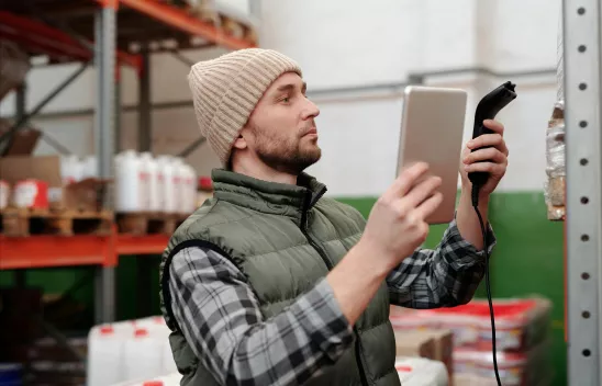 Person mit Mütze und Weste scannt Ware in einem Lager mit Tablet und Handscanner