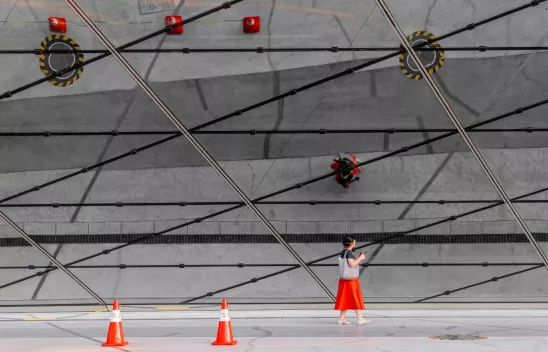 Eine Person mit rotem Rock läuft vor einer Glasfassade, auf der sich die Struktur des Straßenbelags und rote Baustellenkegel spiegeln.
