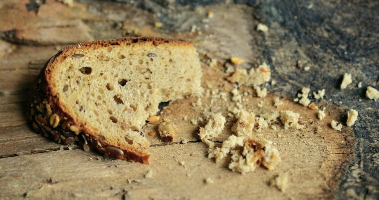 Auf Holz liegt eine halbe Scheibe körniges Brot, welches einmal abgebissen wurde. Daneben liegen Brotkrümel. 
