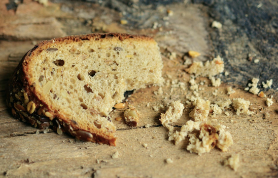 Auf Holz liegt eine halbe Scheibe körniges Brot, welches einmal abgebissen wurde. Daneben liegen Brotkrümel. 