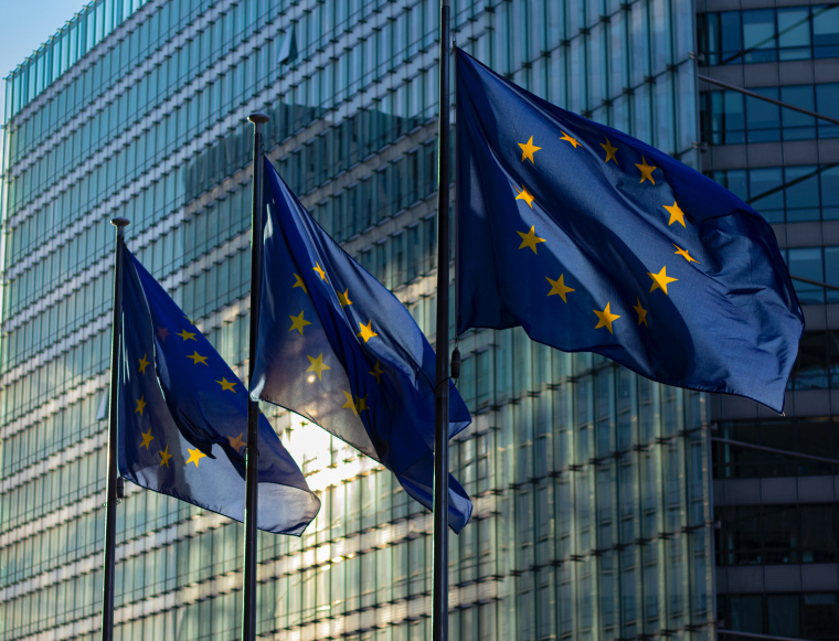 Drei europäische Flaggen (blauer Hintergrund mit einem gelben Sternenkreis) wehen vor einem hohen Glasgebäude.