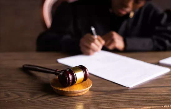 Im Vordergrund liegt ein Gerichtshammer auf einer Holzscheibe auf einem Tisch. Im Hintergrund sieht man, wie eine Person, die am Tisch sitzt, etwas auf ein Blatt Papier schreibt.