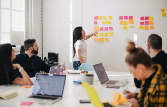 Eine Person steht vor einem Whiteboard, an dem mehrere Post-Its befestigt sind, im Vordergrund sitzen mehrere Personen mit Laptops an einem Tisch