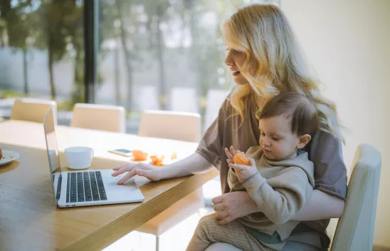 Eine Frau sitzt mit Kind im auf dem Schoß an einem Tisch und arbeitet mit einem Laptop. Das Kind isst eine Mandarine.