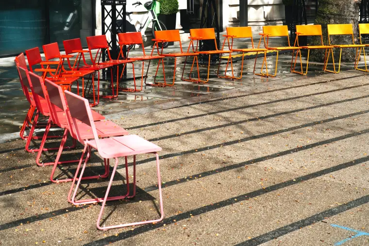 Stühle in den Farben gelb, orange und rot, stehen im Halbkreis auf einem öffentlichen Platz.