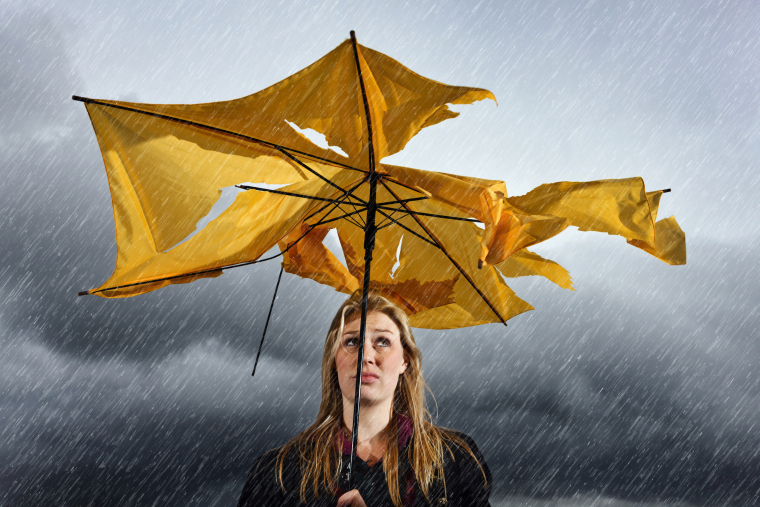 Eine Frau steht im Regen unter grauen Wolken unter einem zerrissenen gelben Regenschirm und blickt hinauf. 