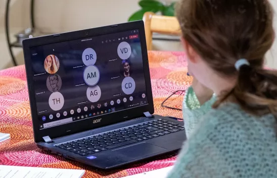 Eine Person sitzt an einem Laptop, auf dem Bildschirm werden Kacheln einer Videokonferenz angezeigt