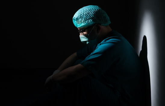 Ein Pfleger mit Kittel, Haarhaube und Maske sitzt in einem dunklen Raum und schaut nach unten. Licht scheint auf ihn. 