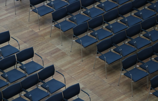 Das Bild zeigt eine große Anzahl leerer, dunkelblauer Stühle, die ordentlich in Reihen auf einem Holzboden angeordnet sind.
