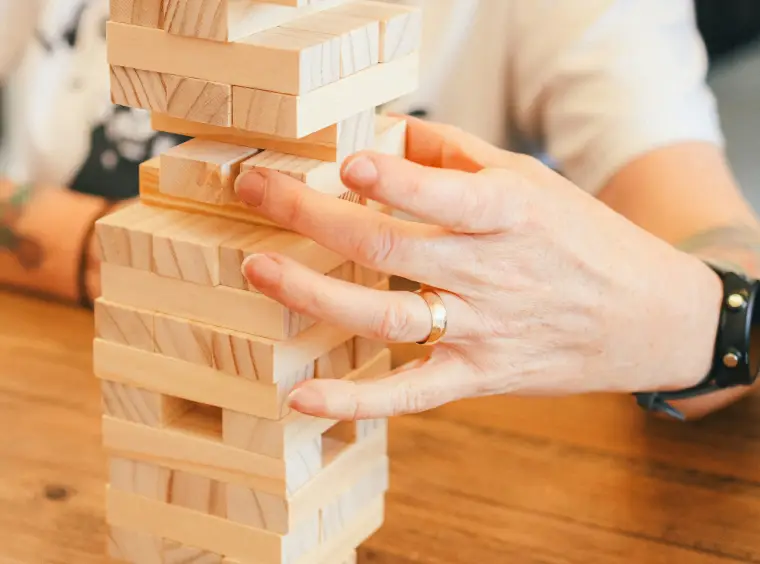 Eine Hand zieht einen Holzbaustein aus einem Turm aus gestapelten Holzbausteinen.