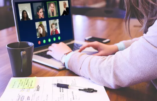 Eine Person sitzt an einem Laptop, auf dem Bildschirm werden Kacheln einer Videokonferenz angezeigt. Neben dem Laptop sind eine Tasse und ein Blatt Papier mit Stift zu sehen.