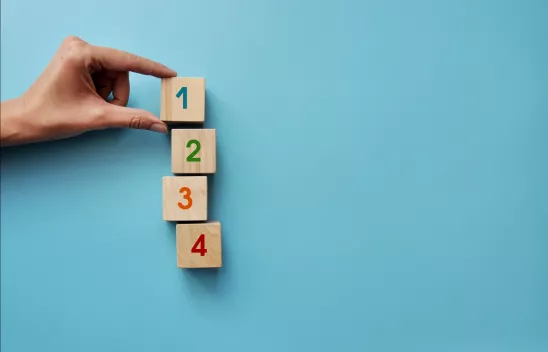 Auf hellblauem Hintergrund liegen vier Würfeln mit den Zahlen von eins bis vier farblich beschriftet. Eine hand hält mit zwei Fingern den ersten Würfel mit der Eins fest. Auf hellblauem Hintergrund liegen vier Würfeln mit den Zahlen von eins bis vier farblich beschriftet. Eine hand hält mit zwei Fingern den ersten Würfel mit der Eins fest.