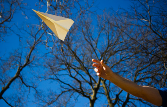 Ein ausgestreckter Arm lässt einen Papierflieger vor blauem Himmel und blätterlosen Bäumen fliegen.