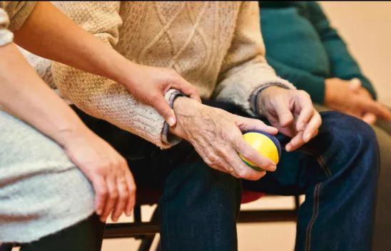 Blick auf zwei Personen, die nebeneinander sitzen. Eine jüngere Person hält die Hand einer älteren Person, in ihrer Hand befindet sich ein kleiner Ball.