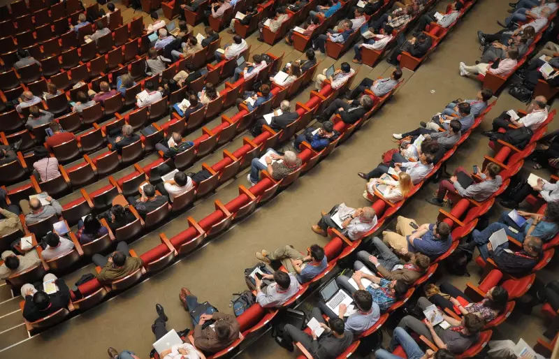 Vogelperspektive auf einen gut besuchten Konferenzsaal mit zahlreichen Personen, die in roten Sitzreihen einer Veranstaltung folgen