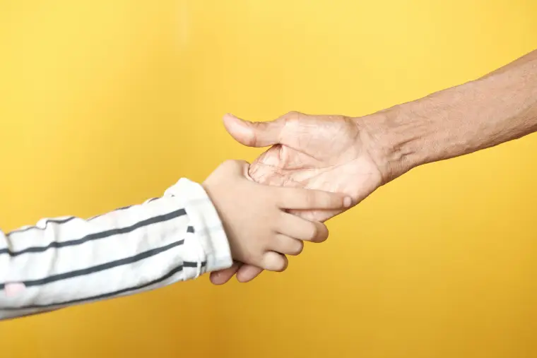 Ein Kind hält die Hand eines älteren Menschen vor einem gelben Hintergrund.