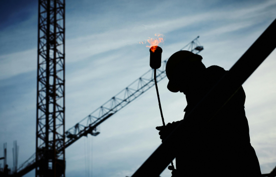 Silhouette eines Bauarbeiters mit Schutzhelm und Gasbrenner vor einem Kran im Hintergrund
