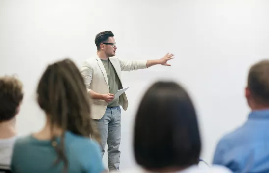 Eine Person steht vorne und präsentiert etwas, im Vordergrund sind verschiedene andere Menschen zu sehen, die dem Vortrag folgen.