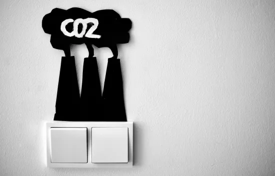 Zwei Lichtschalter nebeneinander. Darüber sind aus Papier drei Kraftwerktürme, die eine Wolke produzieren, in der "CO2" steht.