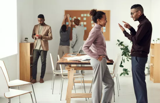 In einem Raum sind mehrere Personen zu sehen, zwei Personen beschäftigen sich mit einer Pinnwand, im Vordergrund unterhalten sich zwei Personen