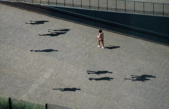 Person auf grauem Asphalt gehend. Ringsherum mehrere menschliche Schatten, zu denen aber keine Personen sichtbar sind.