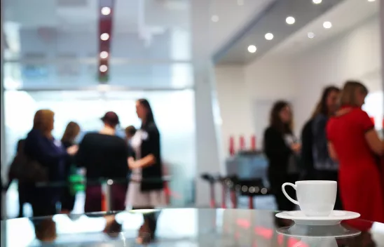 Im Vordergrund ein Stehtisch, auf dem eine kleine Kaffeetasse zu sehen ist, im Hintergrund sind Personen in Gruppen zu sehen, die miteinander sprechen
