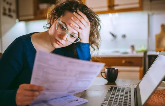 Eine Frau hat ihren Kopf auf einer Hand aufgestützt und sitzt vor einem Laptop. Sie hat ein Formular oder eine Rechnung in der anderen Hand und schaut darauf. Eine Frau hat ihren Kopf auf einer Hand aufgestützt und sitzt vor einem Laptop. Sie hat ein Formular oder eine Rechnung in der anderen Hand und schaut darauf.