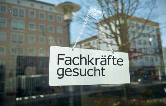 Auf einer Glasscheibe hängt ein Schild mit der Aufschrift "Fachkräfte gesucht". Es spiegeln sich Häuser im Hintergrund in der Scheibe.