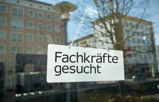 Auf einer Glasscheibe hängt ein Schild mit der Aufschrift "Fachkräfte gesucht". Es spiegeln sich Häuser im Hintergrund in der Scheibe. Auf einer Glasscheibe hängt ein Schild mit der Aufschrift "Fachkräfte gesucht". Es spiegeln sich Häuser im Hintergrund in der Scheibe.