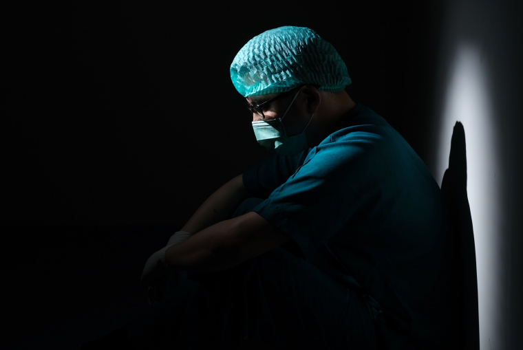 Ein Pfleger mit Kittel, Haarhaube und Maske sitzt in einem dunklen Raum und schaut nach unten. Licht scheint auf ihn. 